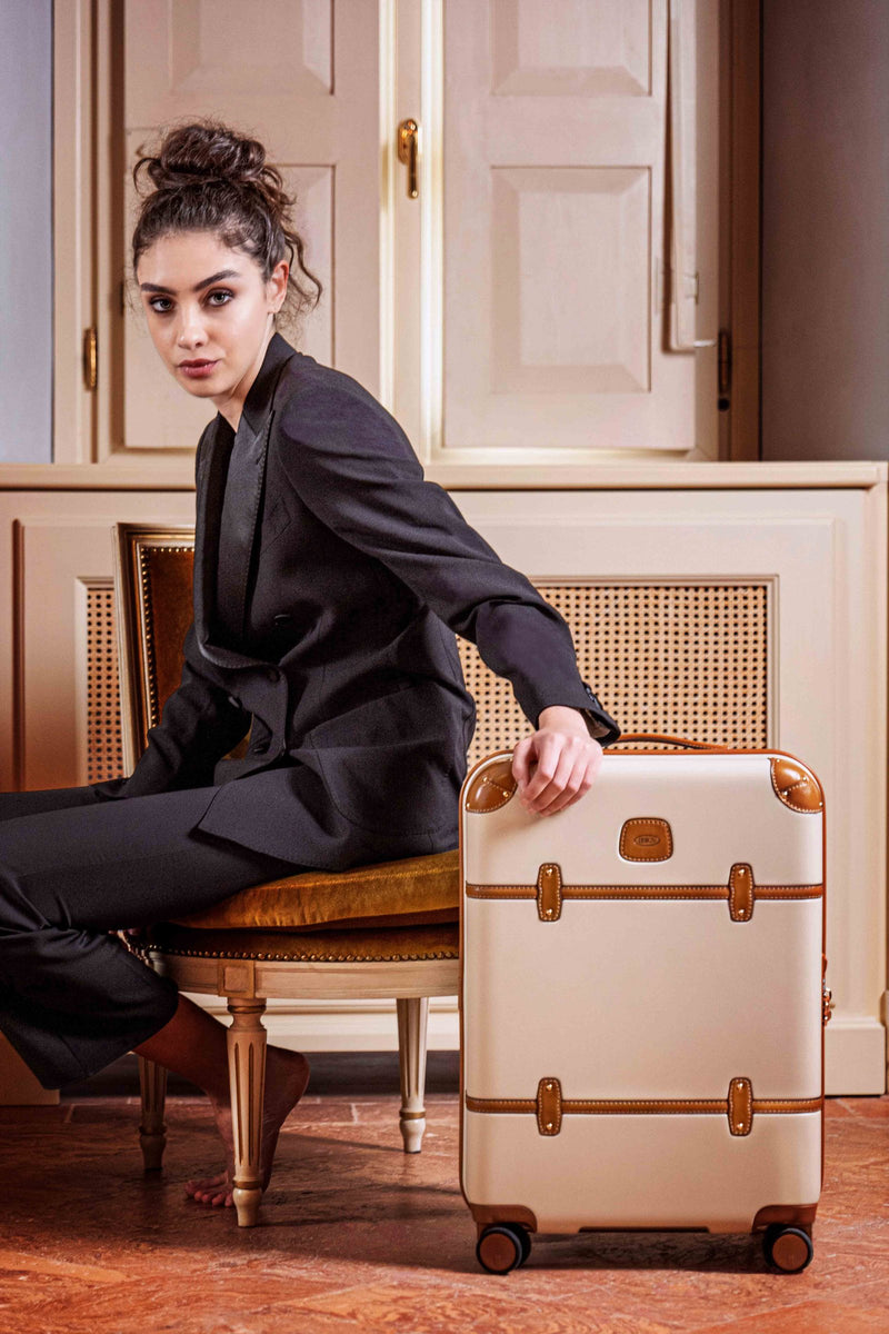 Woman in a dark suit seated beside a cream Bric's Bellagio carry-on spinner in a classic interior