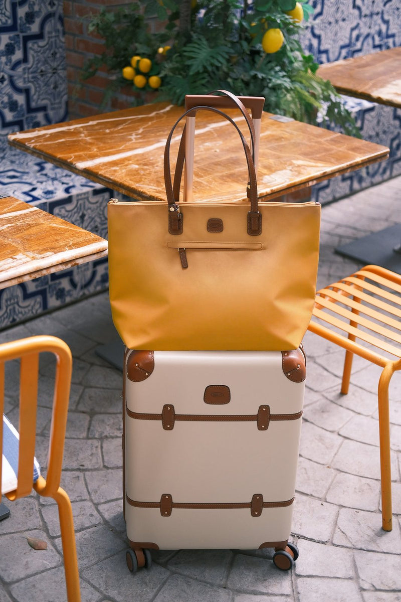 Cream Bric's Bellagio carry-on spinner with a tan tote resting on top, beside a cafe table