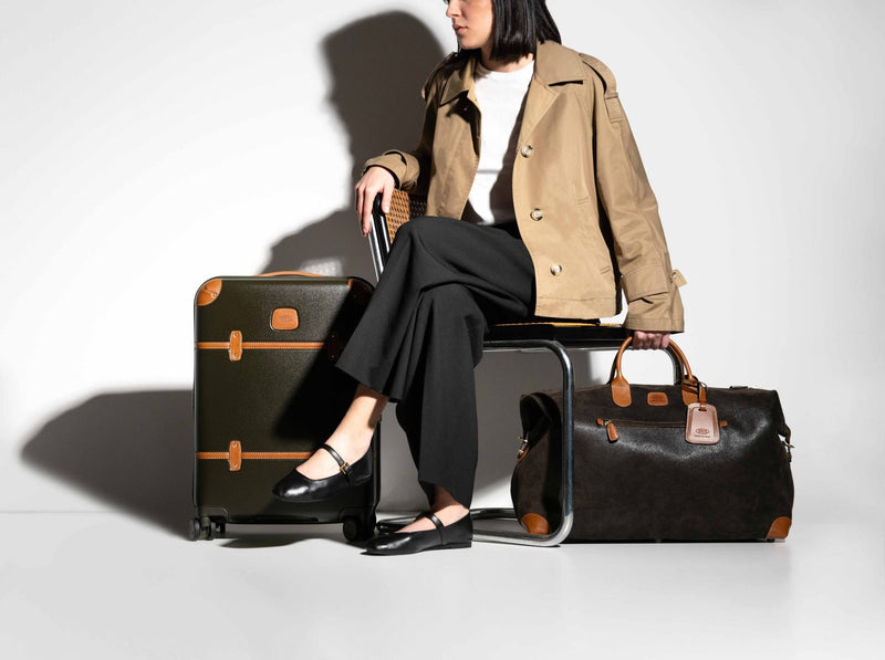 Woman seated beside an olive Bric's Bellagio carry-on spinner and a leather-trimmed duffle