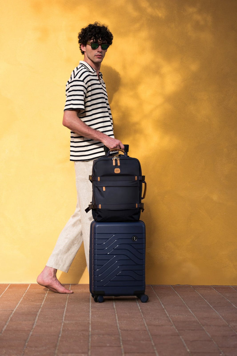 Man in a striped shirt against a yellow wall with an Ocean Blue Bric's B|Y Ulisse carry-on and a bag stacked on top