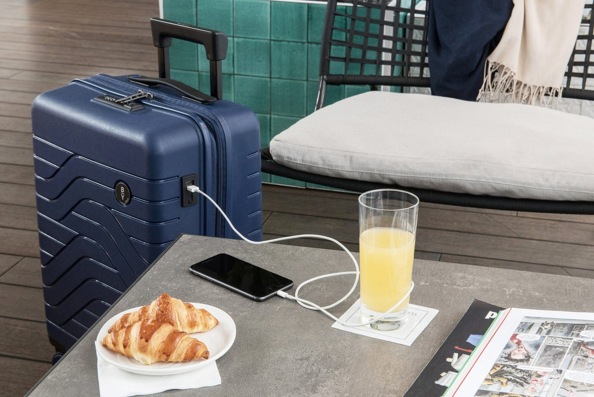 Ocean Blue Bric's B|Y Ulisse carry-on charging a phone on a terrace table beside croissants and juice