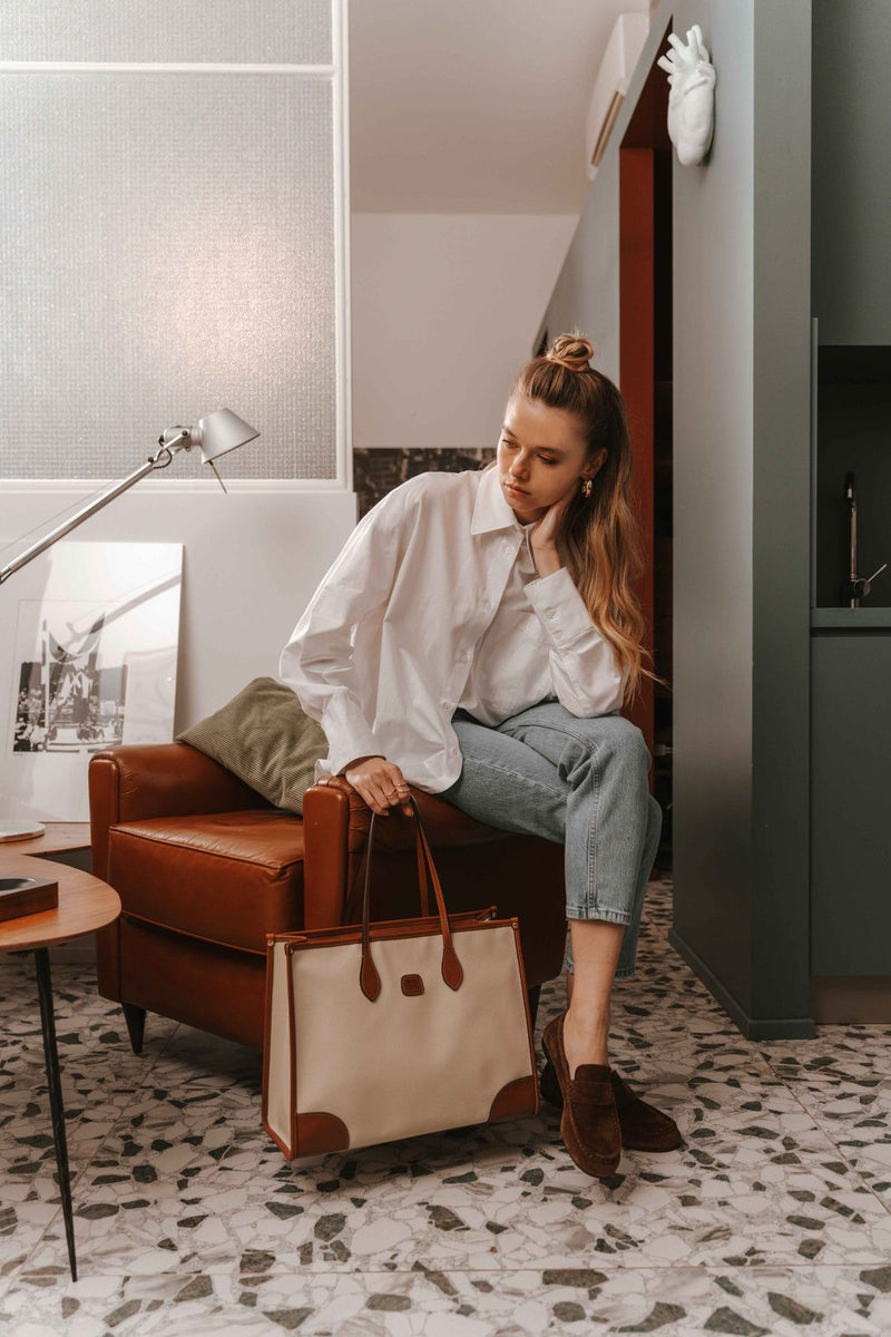 Woman seated in a lounge chair with a cream Bric's Firenze laptop tote at her feet