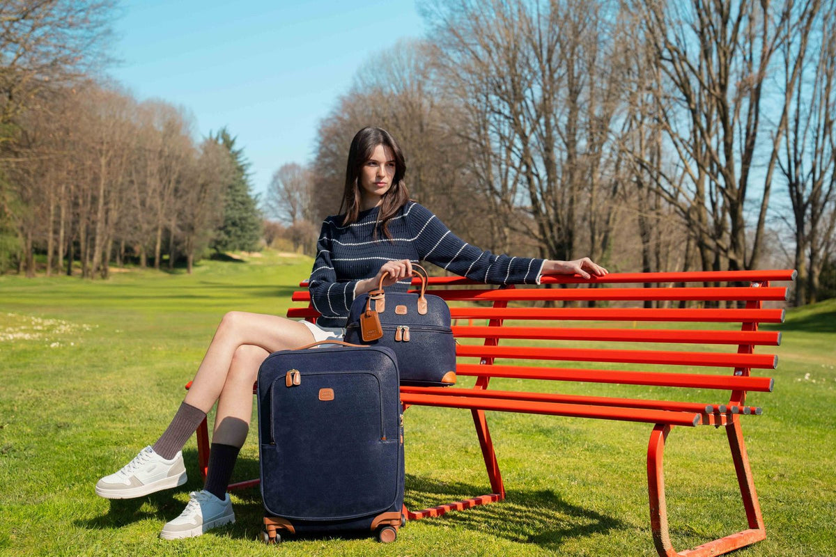 Woman on a red park bench with a blue Bric's Life Compound carry-on spinner on the grass beside her