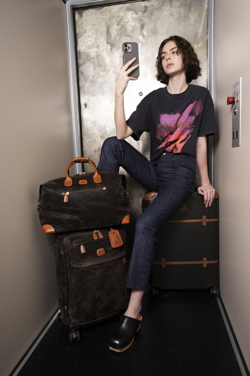 Olive Bric's Life cargo duffle stacked on a spinner in an elevator, beside a woman taking a selfie