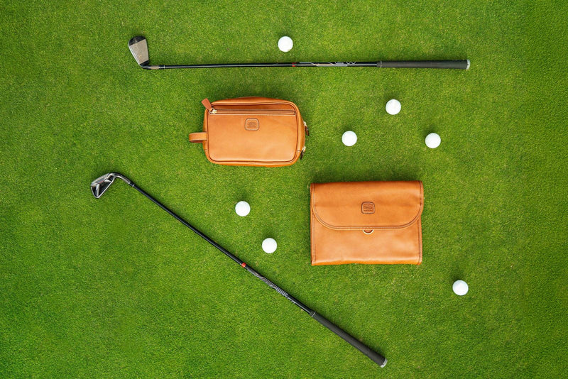 Two cognac leather Bric's Life Pelle toiletry bags on a golf green with clubs and golf balls