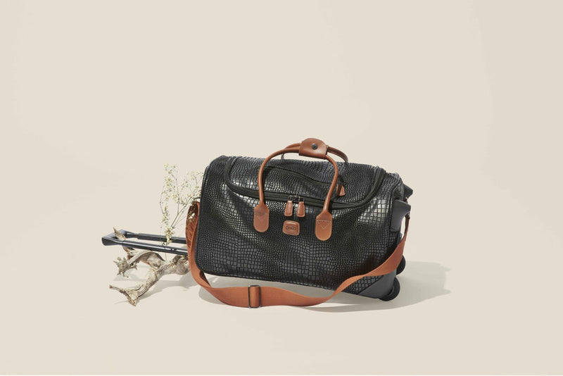 Black Bric's My Safari rolling duffle with tan leather handles, set beside driftwood and dried flowers