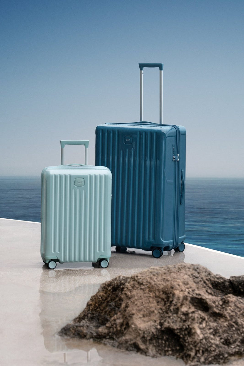 Bric's Positano spinner in Sea Green standing on a seaside terrace beside a smaller pale case