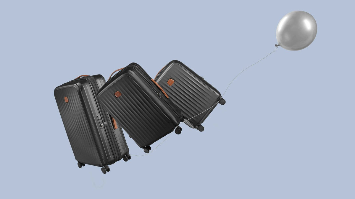Three black Bric's Taormina carry-ons drifting on a pale blue background, tied to a silver balloon