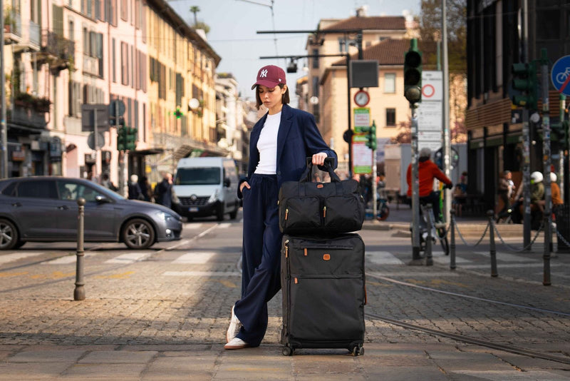 Woman on a city street resting her hand on a black Bric's soft-side spinner with a black duffle on top
