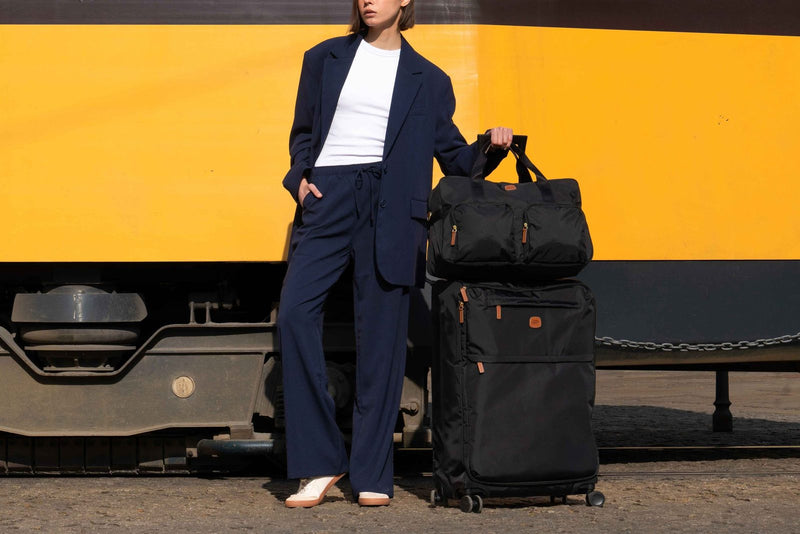 Woman in a navy suit beside a black Bric's X-Collection boarding duffle stacked on a black spinner