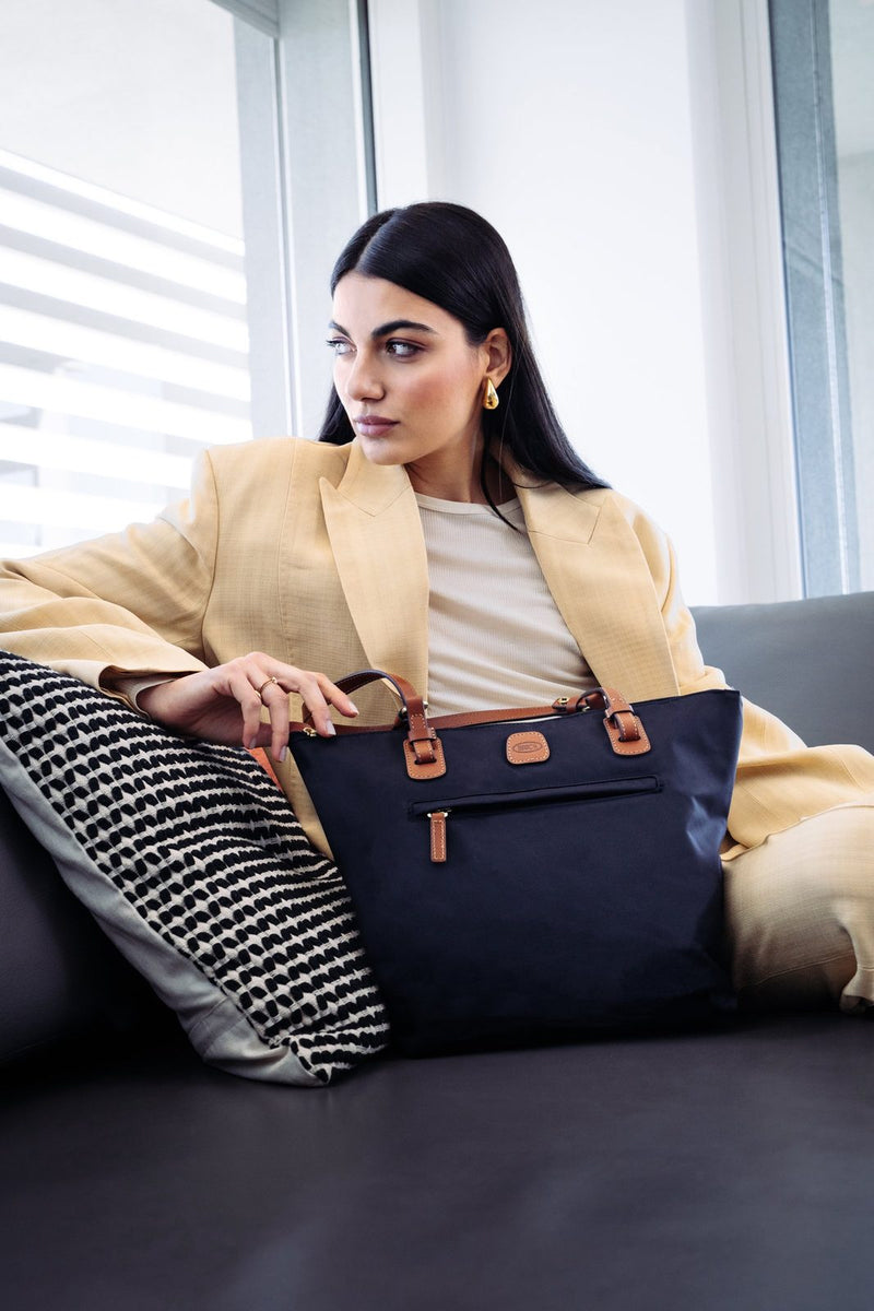 Woman in a cream suit on a sofa with a navy Bric's X-Collection Sportina bag beside her