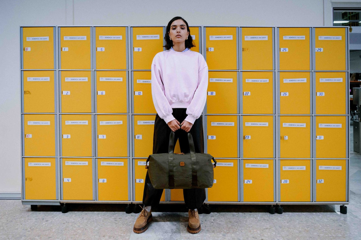 Woman holding an olive Bric's X-Collection folding duffle in front of a wall of yellow lockers