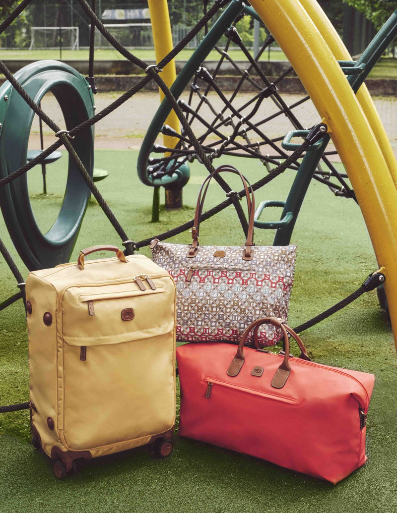 Poppy Bric's X-Collection deluxe duffle on a playground lawn beside a yellow spinner and a patterned tote